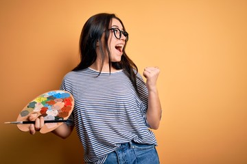 Young brunette artist woman painting holding painter brush and palette over yellow background pointing and showing with thumb up to the side with happy face smiling