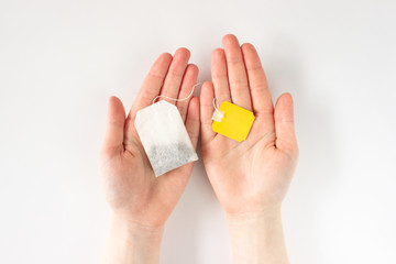 White tea bag with yellow label in female hands on white background top view in flat lay style. Tea bag or loose leaf tea, waste and recycle concept. Stock photo