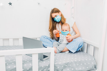 A young mother in home pajamas and a medical protective mask on her face, with a small child...