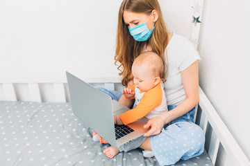 A young mother in home pajamas and a medical protective mask on her face, with a small child...
