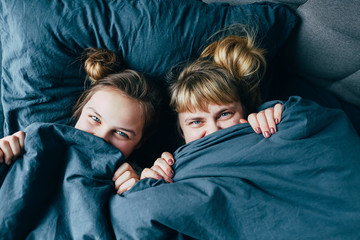 Portrait of light hair long hair mother and daughter under the duvet together in soft morning light on blue linen bed. Concept of happy family living, relaxation, comfort, fun. Play hide and seek - 341072079