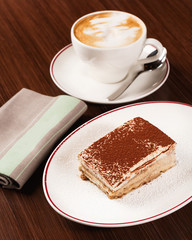 Tiramisu. Original recipe of Italian dessert with cacao powder on top with cup of cappuccino. Authentic Italian cuisine. Wooden table
