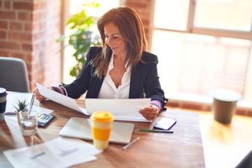 Middle age beautiful businesswoman smiling happy and confident. Sitting on chair working in a desk reading documents at the office