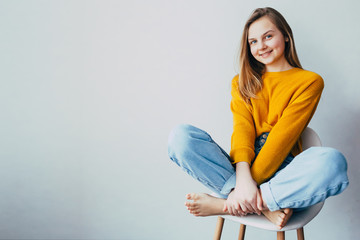 teenage girl looks at camera .sitting on white modern chair with legs up. Beautiful girl in yellow sweater and blue jeans in home interior. Trendy casual outfit. place for text - 341072420