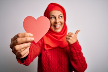 Middle age brunette business woman wearing traditional muslim hijab holding paper heart pointing and showing with thumb up to the side with happy face smiling