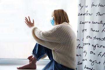 Concept of coronavirus covid-19 quarantine or pandemic prevention. Young attractive blonde girl wearing medical protective face mask during flu virus, forced to stay at home, looking out of window.
