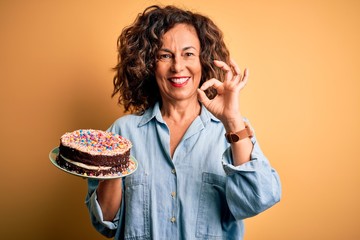 Middle age beautiful woman holding cake standing over isolated yellow background doing ok sign with fingers, excellent symbol