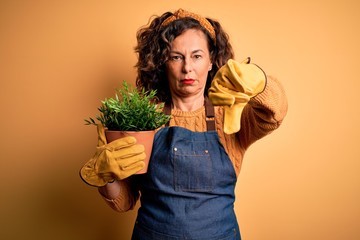 Middle age gardener woman wearing apron holding plant pot over isolated yellow background with...