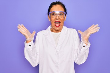 Naklejka premium Middle age senior scientist woman wearing coat and laboratory glasses over purple background celebrating crazy and amazed for success with arms raised and open eyes screaming excited. Winner concept