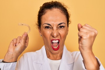Middle age senior dentist woman holding clear aligner for teeth correction annoyed and frustrated...
