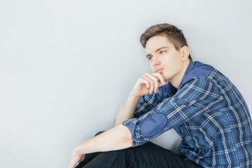 A young man is sitting near a light gray wall. Pensive young man in a blue shirt