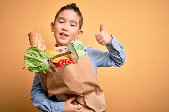 Adorable toddler holding paper bag with food standing over isolated yellow background