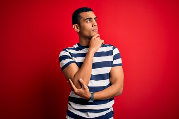 Handsome african american man wearing casual striped t-shirt standing over red background with hand on chin thinking about question, pensive expression. Smiling with thoughtful face. Doubt concept.