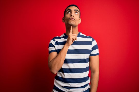 Handsome african american man wearing casual striped t-shirt standing over red background Thinking concentrated about doubt with finger on chin and looking up wondering