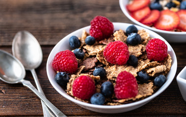Granola with blueberries and raspberries in white bowl in dark wooden desk.