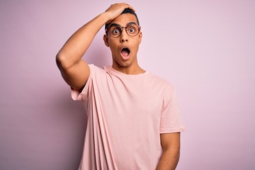 Handsome african american man wearing casual t-shirt and glasses over pink background surprised...