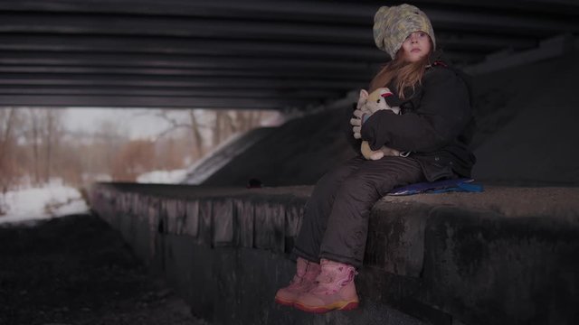 A helpless scared little homeless girl in a holey knitted multi-colored hat sits in the winter under the bridge. The child hugs a single beige soft toy