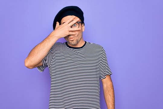 Handsome man with blue eyes wearing striped t-shirt and french beret over purple background peeking in shock covering face and eyes with hand, looking through fingers with embarrassed expression.
