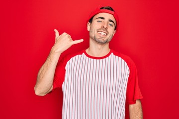 Young handsome sporty man with blue eyes wearing striped baseball t-shirt and cap smiling doing phone gesture with hand and fingers like talking on the telephone. Communicating concepts.