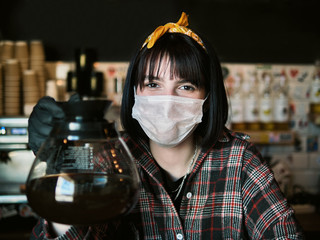 Concept of biohazard security and social responsibility in coronavirus epidemic measures to contain virus spread: young barista girl in medical mask and medical gloves holding coffee jug in city cafe