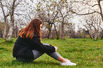Sad girl sitting alone on grass in park vintage style toned photo. Loneliness depression concept