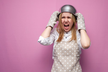 young unhappy girl cook in kitchen clothes with a pan on her head screams on a colored background, woman housewife with kitchen items in stress