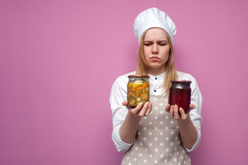 young thoughtful girl cook in kitchen clothes holds canned food and thinks on a colored background, woman housewife with food blanks