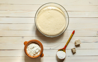 yeast dough in a bowl, a ceramic pot with flour and a wooden painted spoon, top view