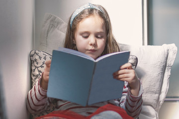 Caucasian girl reading a book with some tales and stories sitting on cushions. At home schooling. Gifted children concept.