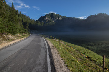 Obraz premium A small, light morning fog rising above the meadow by the asphalt road. Dolomite, Italy.