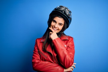 Young beautiful brunette motorcycliste woman wearing motorcycle helmet and jacket looking confident at the camera smiling with crossed arms and hand raised on chin. Thinking positive.