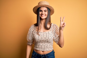 Young beautiful brunette woman wearing t-shirt and summer hat over yellow background smiling with...