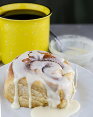 Cinnamon roll on a white plate with cup of coffee and bowl of glaze in the background.