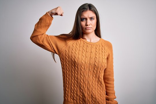 Young beautiful woman with blue eyes wearing casual sweater standing over white background Strong person showing arm muscle, confident and proud of power
