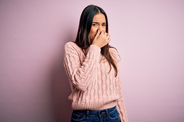 Young beautiful brunette woman wearing casual sweater over isolated pink background smelling something stinky and disgusting, intolerable smell, holding breath with fingers on nose. Bad smell