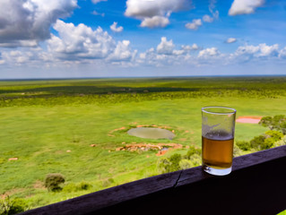 A glass of beer stands on a wooden fence. In the background is a wonderful view of Tsavo East National Park in Kenya.