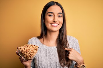 Young beautiful brunette woman holding bowl with peanuts over isolated yellow background with surprise face pointing finger to himself