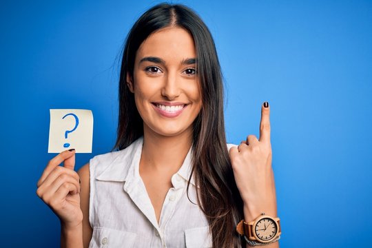 Young beautiful brunette woman holding paper with question mark symbol message surprised with an idea or question pointing finger with happy face, number one