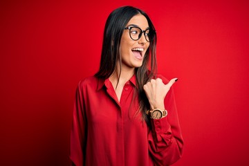 Young beautiful brunette woman wearing casual shirt and glasses over red background smiling with...