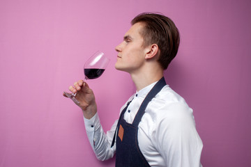 young sommelier guy checks the smell of red wine on a colored background, an expert on wine in a white shirt and apron holds a glass, the waiter tastes the wine