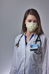 Masked healthcare worker smiling and posing under yellow mask against a grey background. Stethoscope, white coat, and PPE visible in portrait
