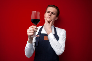 professional thoughtful sommelier holds a glass of red wine and thinks against a red background, the guy the waiter looks at the wine - obrazy, fototapety, plakaty