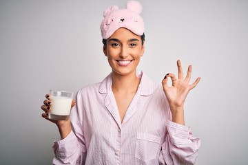 Young beautiful woman wearing pajama and sleep mask drinking glass of healthy milk doing ok sign with fingers, excellent symbol