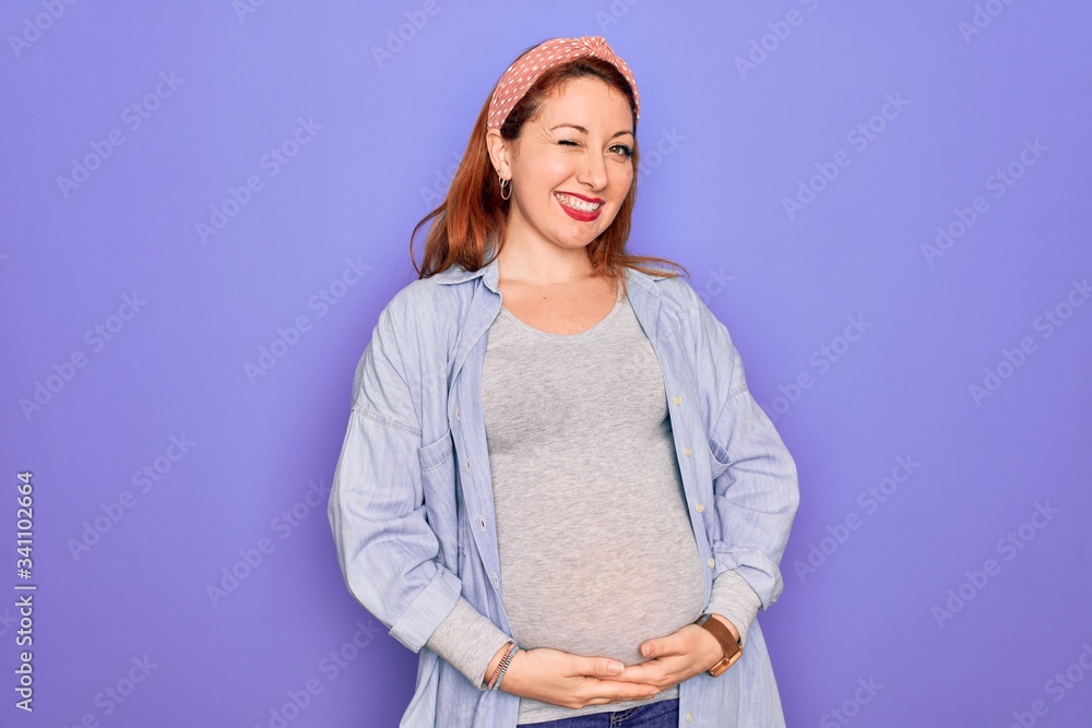 Sticker young beautiful redhead pregnant woman expecting baby over isolated purple background winking lookin