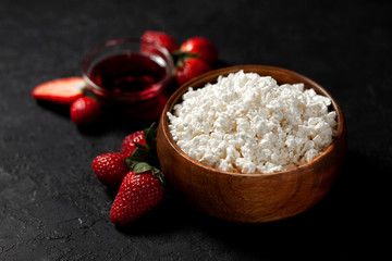 tasty homemade cottage cheese in a wooden plate with strawberries and jam on a dark background, healthy breakfast on a black table