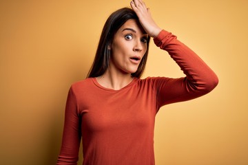Young beautiful brunette woman wearing casual t-shirt standing over yellow background surprised...