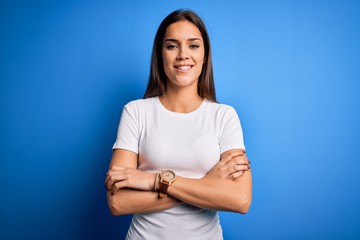 Obraz premium Young beautiful brunette woman wearing white casual t-shirt standing over blue background happy face smiling with crossed arms looking at the camera. Positive person.
