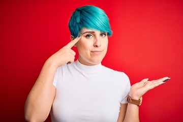 Young beautiful woman with blue fashion hair wearing casual t-shirt over red background confused and annoyed with open palm showing copy space and pointing finger to forehead. Think about it.