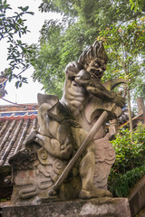 Fototapeta na wymiar Fengdu, China - May 8, 2010: Ghost City, historic sanctuary. Closeup of brown stone hatchet holding man-like angry monster statue on wall under green foliage and silver sky.