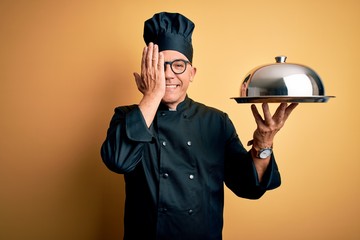 Middle age handsome grey-haired waiter man wearing cooker uniform and hat holding tray covering one eye with hand, confident smile on face and surprise emotion.